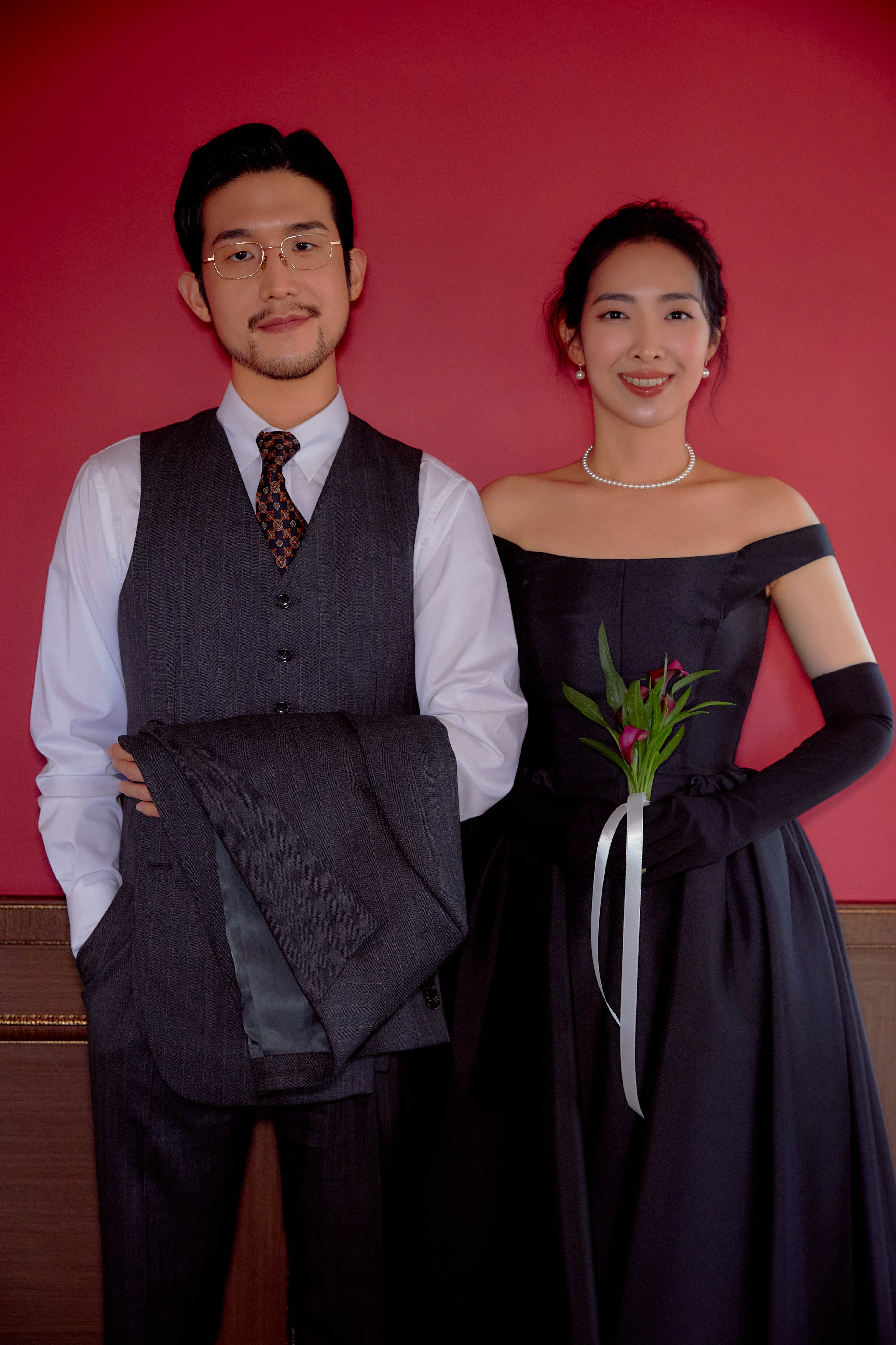 웨딩 사진 메인. 와인색 벽, 팔짱 끼고 있으며 신부는 부케를 들고 있다.